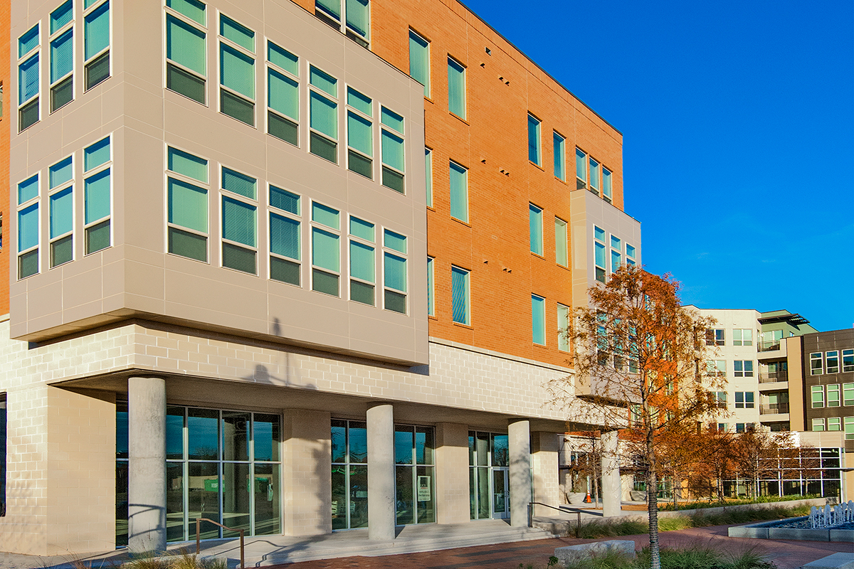 Modern multi-story building with large windows, a brick and beige exterior, concrete columns, and a small tree in front, set against a clear blue sky.