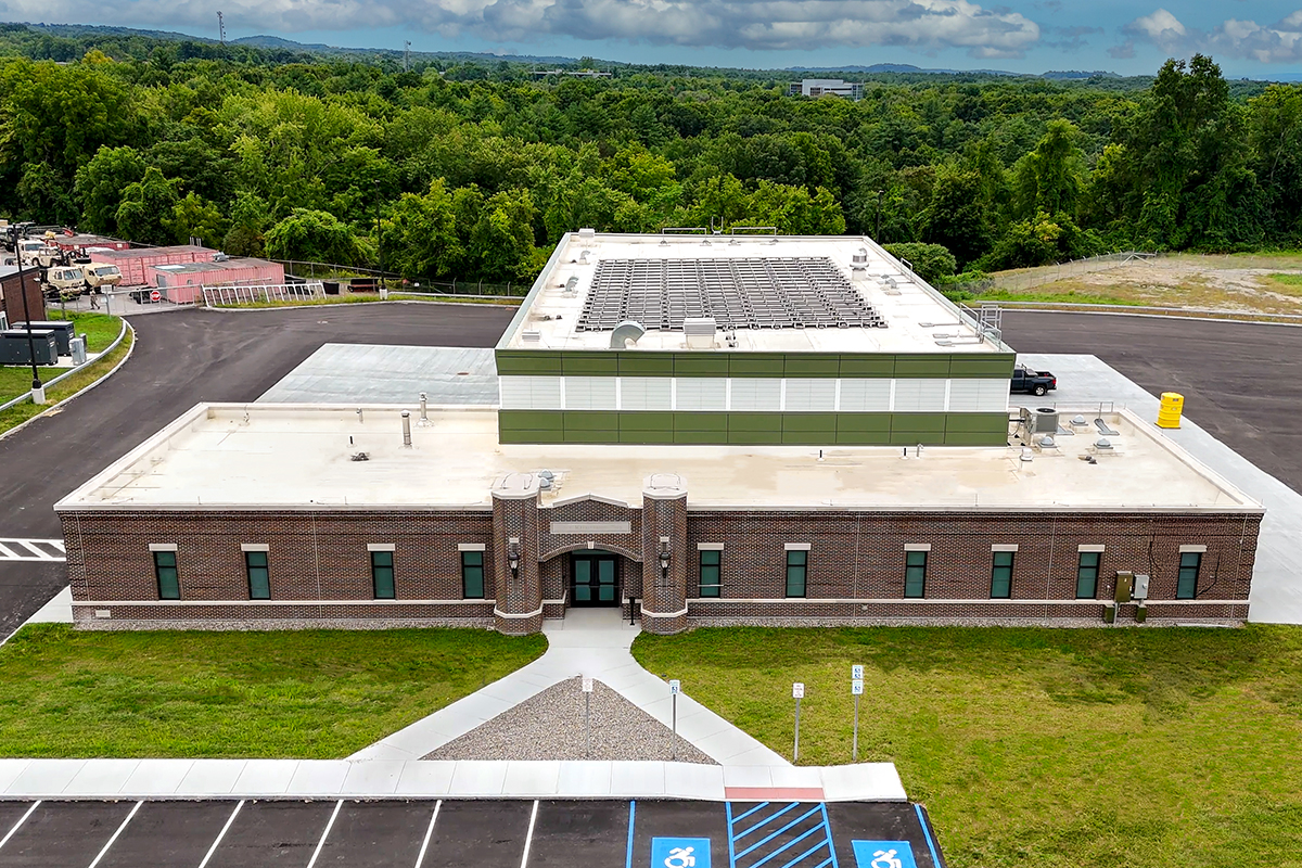 A single-story brick building with a flat white roof, surrounded by greenery. The entrance has a small walkway and accessible parking spaces in front. Trees and a partly cloudy sky are in the background.