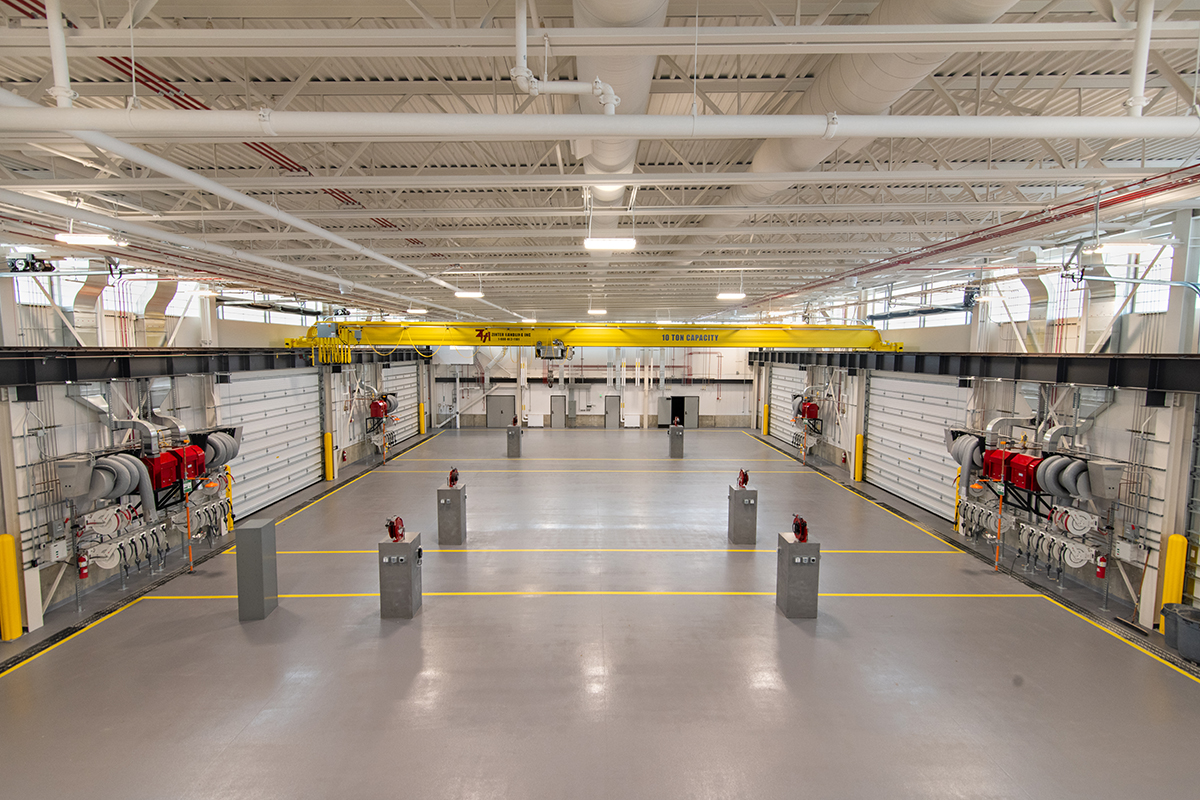 A spacious, brightly lit industrial facility features a yellow overhead crane, red equipment along the walls, ventilation ducts, and multiple concrete pillars spaced across the shiny, clean floor.