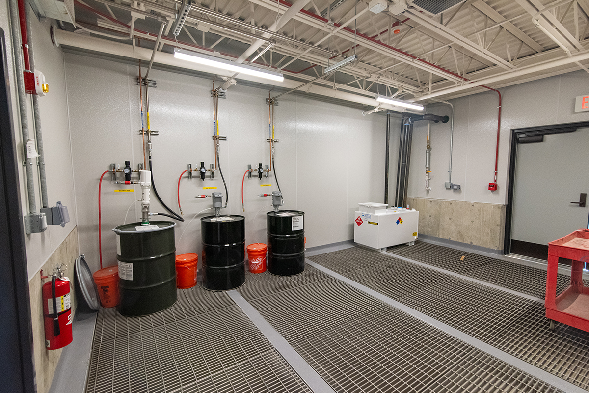 Industrial room with three large black barrels, orange buckets, a white hazardous materials box, metal grates on the floor, and visible pipes and wires on the ceiling and walls.