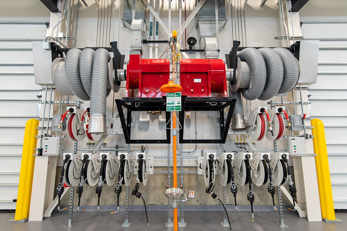 Industrial equipment with red and gray machinery, large hoses, and multiple retractable reels mounted on a wall. An emergency shower with a yellow pull handle is centered in front of the equipment.