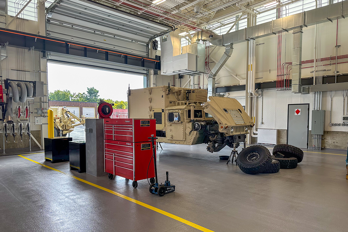 Military vehicle in a maintenance garage with several tires removed, stacked nearby. A red tool chest, jack, and various tools are visible; the large garage door is open, letting in natural light.