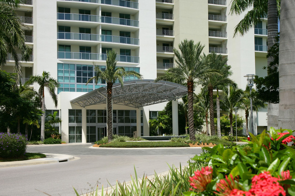A modern hotel entrance with a metal canopy, palm trees, and well-kept landscaping, including red flowers in the foreground, under clear skies.