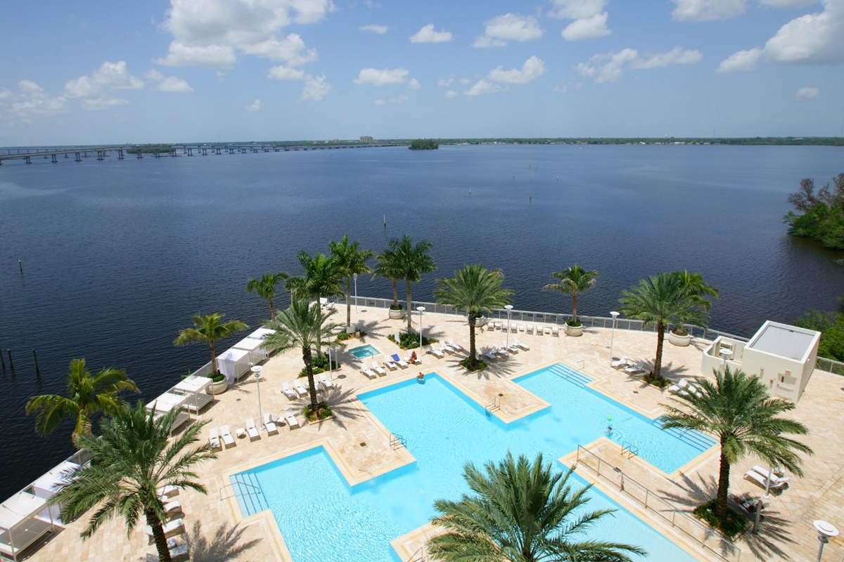 A large pool with palm trees overlooks a calm waterfront and distant bridge under a partly cloudy sky. Lounge chairs line the pool area, creating a relaxing resort atmosphere.