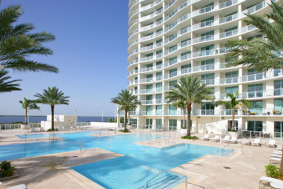 A modern high-rise building with curved balconies overlooks a large, resort-style outdoor pool surrounded by palm trees, lounge chairs, and clear blue sky.