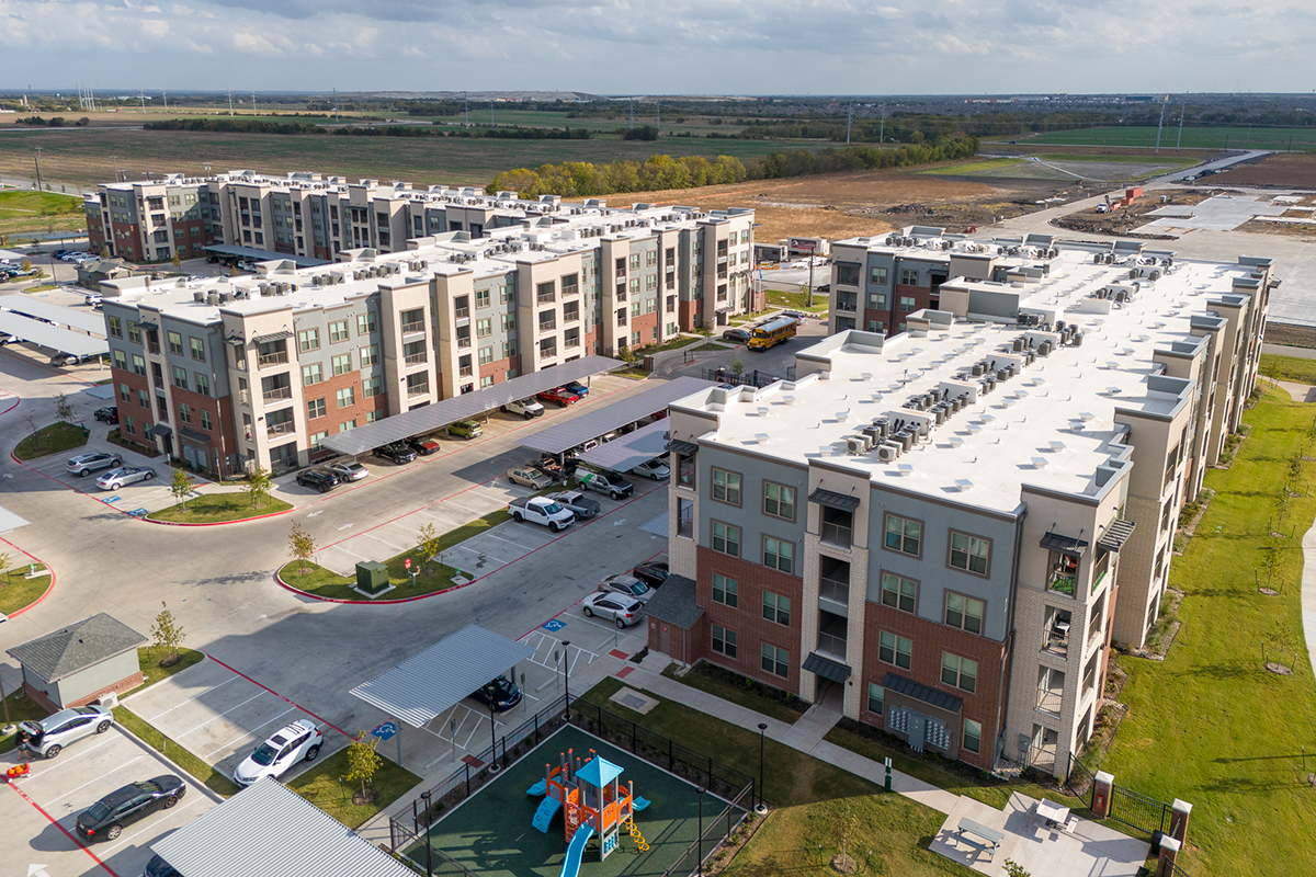 Aerial view of modern apartment buildings with parking lots, cars, and a playground in the foreground, surrounded by open fields and distant roads.