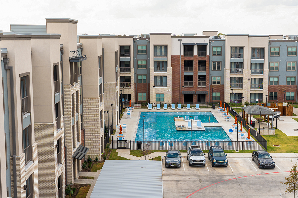 Four-story apartment buildings surround a rectangular outdoor swimming pool with lounge chairs. Several cars are parked in front, and a small covered area sits to the side of the pool.