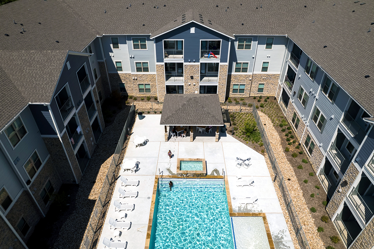 Aerial view of a rectangular swimming pool and hot tub in the courtyard of a modern apartment complex, with lounge chairs and a few people by the water.