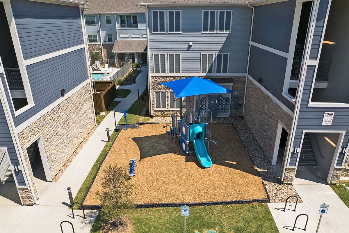 A blue playground with a slide and climbing area sits between two gray apartment buildings, shaded by a blue canopy, with mulch ground and a small tree nearby.
