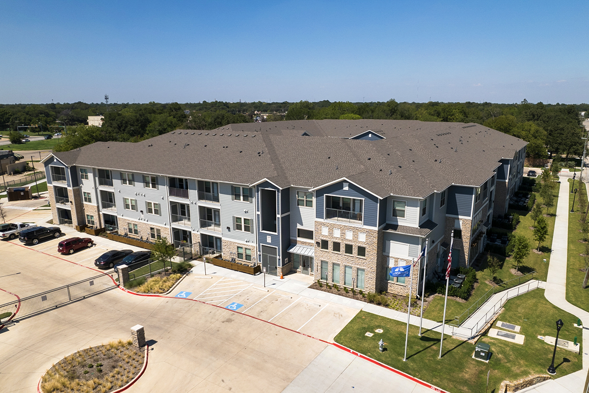 A three-story apartment building with gray siding, several windows, and accessible parking spaces in front, surrounded by greenery under a clear blue sky.