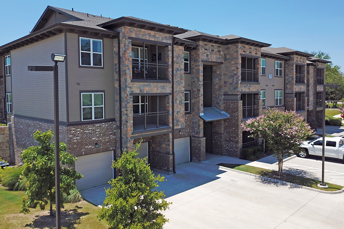 A modern, three-story apartment building with brick and siding exterior, balconies, and garages. Trees and a parking lot are in front, under a clear blue sky.