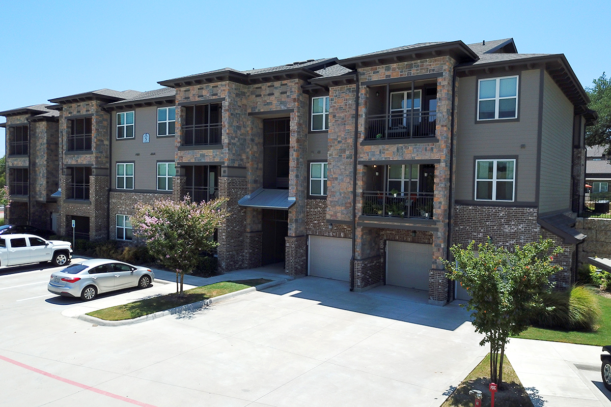 Modern three-story apartment building with stone and brick exterior, balconies, and garages on the ground level. Two cars are parked in front, with small trees and a clear blue sky in the background.