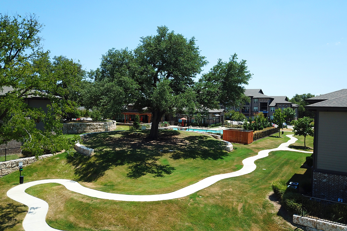 A curving sidewalk winds through a grassy area with large trees, surrounded by apartment buildings. A fenced pool and stone landscaping are visible under a clear blue sky.