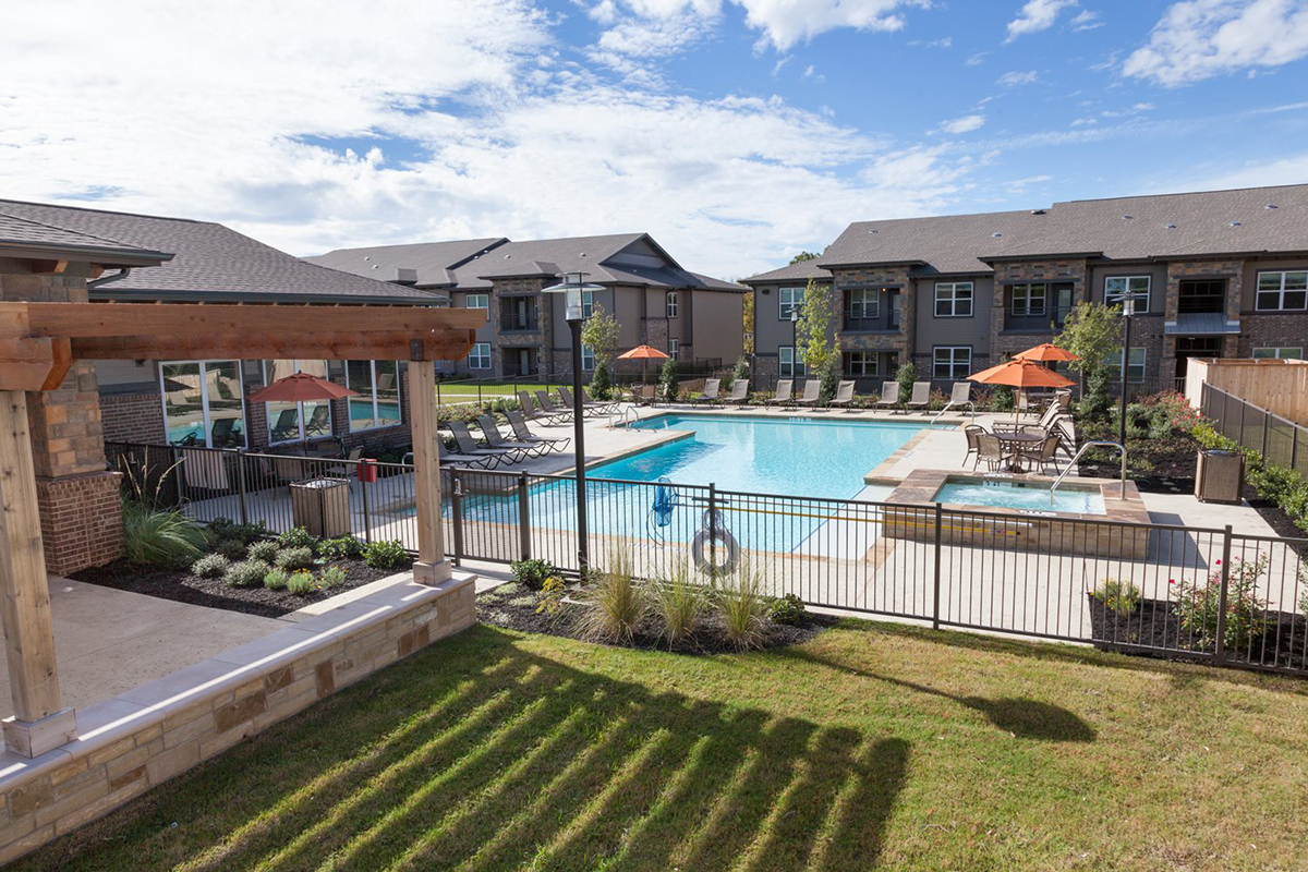 A modern apartment complex courtyard features a fenced outdoor swimming pool, lounge chairs, orange umbrellas, landscaped greenery, and surrounding two-story buildings under a partly cloudy sky.
