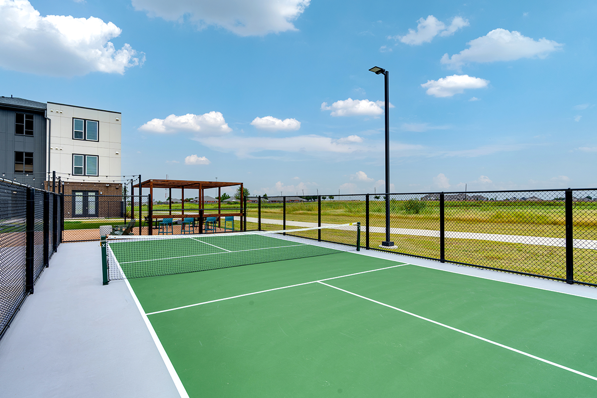 Outdoor pickleball court with a green surface, black fencing, and a net. Nearby, theres a shaded seating area with tables and chairs. Modern building and grassy fields are visible under a blue sky with scattered clouds.