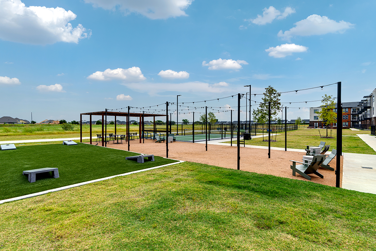 Outdoor community space with string lights, benches, cornhole boards, paved walkways, grassy areas, and nearby apartments under a blue sky with scattered clouds.