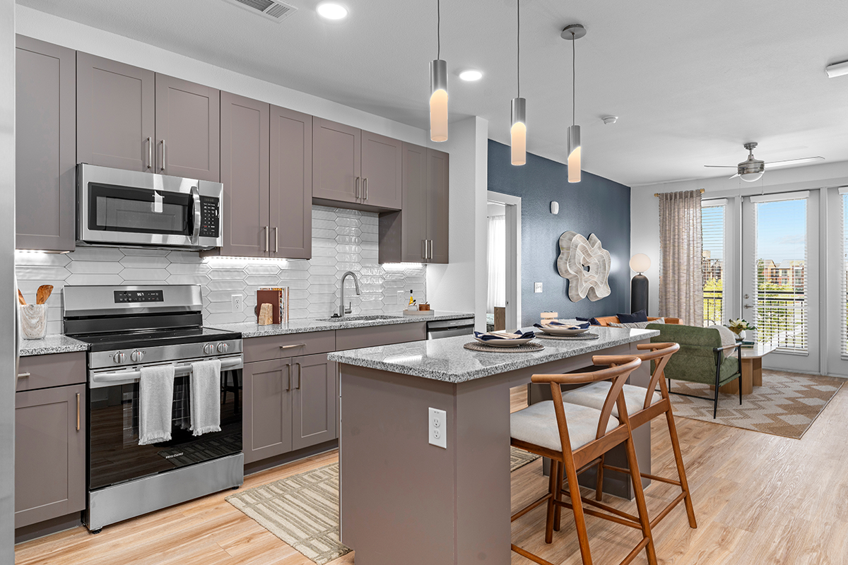 Modern kitchen with gray cabinets, stainless steel appliances, a center island with seating, and pendant lights. The space opens to a living area with large windows, a green sofa, and wooden flooring throughout.
