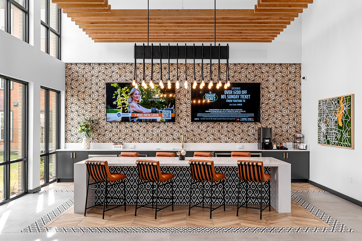 Modern lounge with a long island, five patterned chairs, geometric wall, two TVs, hanging lights, indoor plants, and large windows letting in natural light.