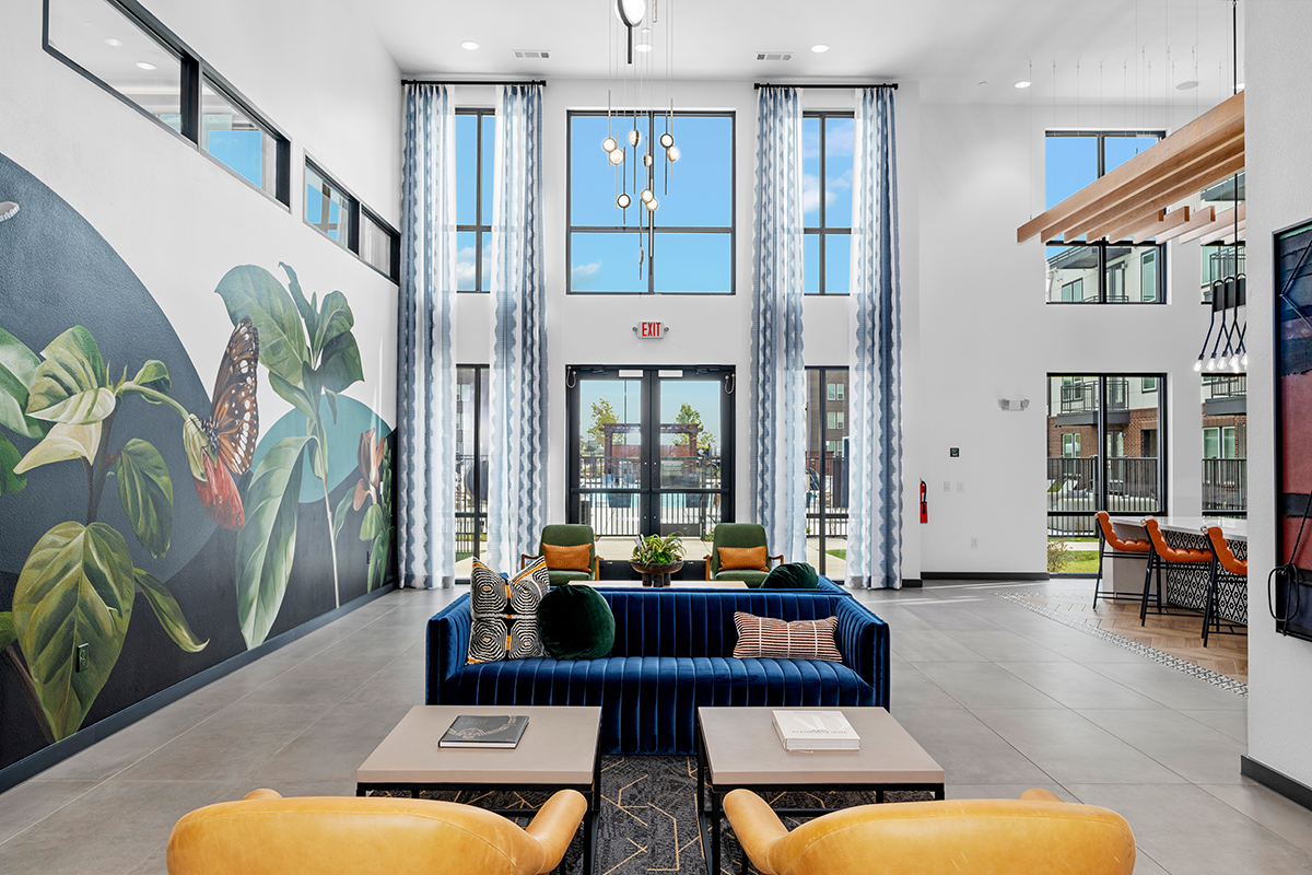 Modern lobby with a blue sofa, yellow chairs, large windows, and a tropical mural on the left wall. Bright natural light fills the space, and tables with books are set in the center.