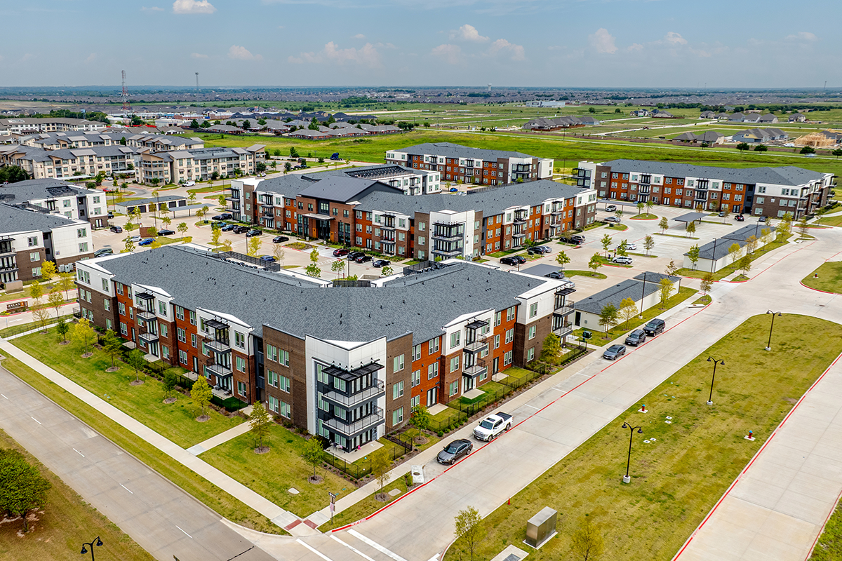 Aerial view of a modern apartment complex with multiple buildings, parking lots, green spaces, and cars parked along roads in a suburban area under a partly cloudy sky.