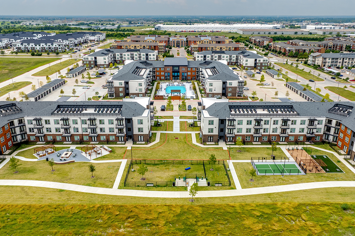 Aerial view of a modern apartment complex with landscaped lawns, walking paths, a central pool area, tennis courts, and picnic spaces, surrounded by parking lots and neighboring buildings.