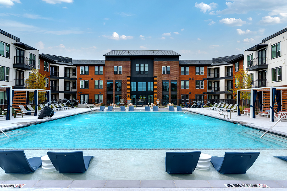 Modern apartment complex with a large, rectangular swimming pool, lounge chairs in the water, and more seating around the pool, surrounded by three-story buildings under a blue sky.