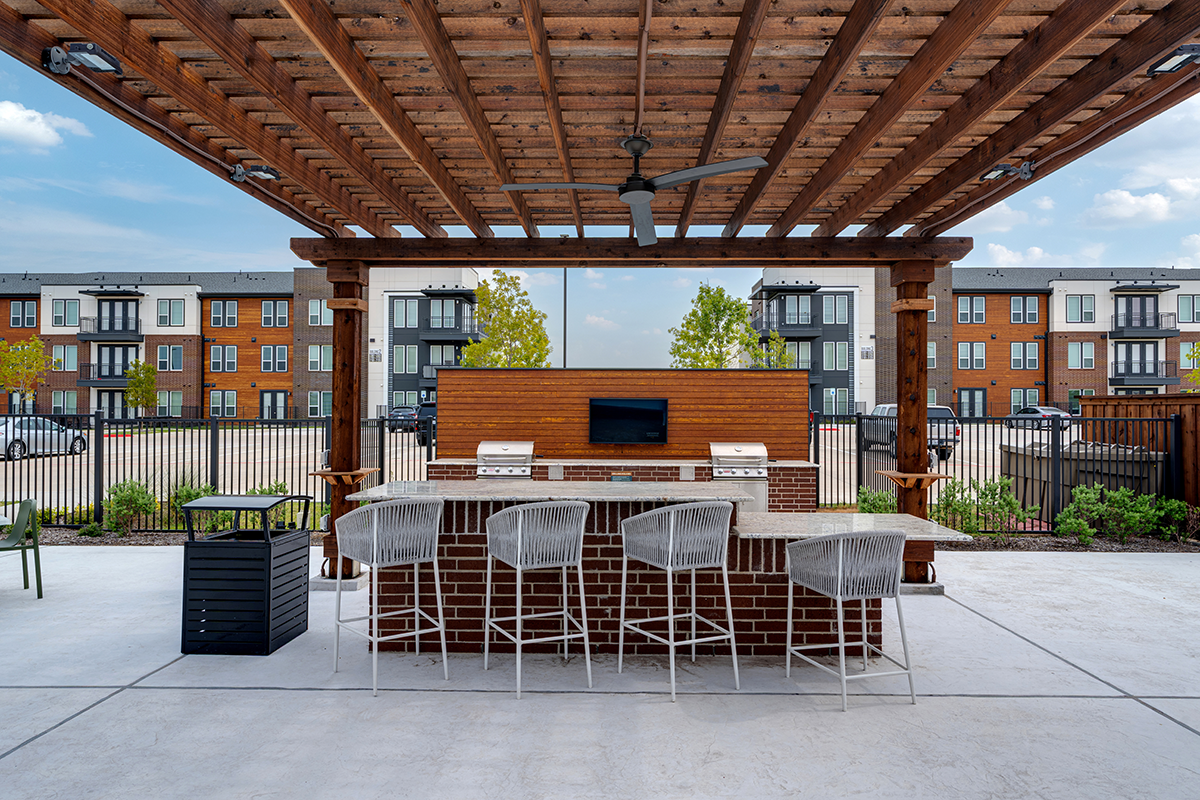 Outdoor kitchen under a wooden pergola with a ceiling fan, grill, TV, and four white barstools. Apartment buildings and parked cars are visible in the background.