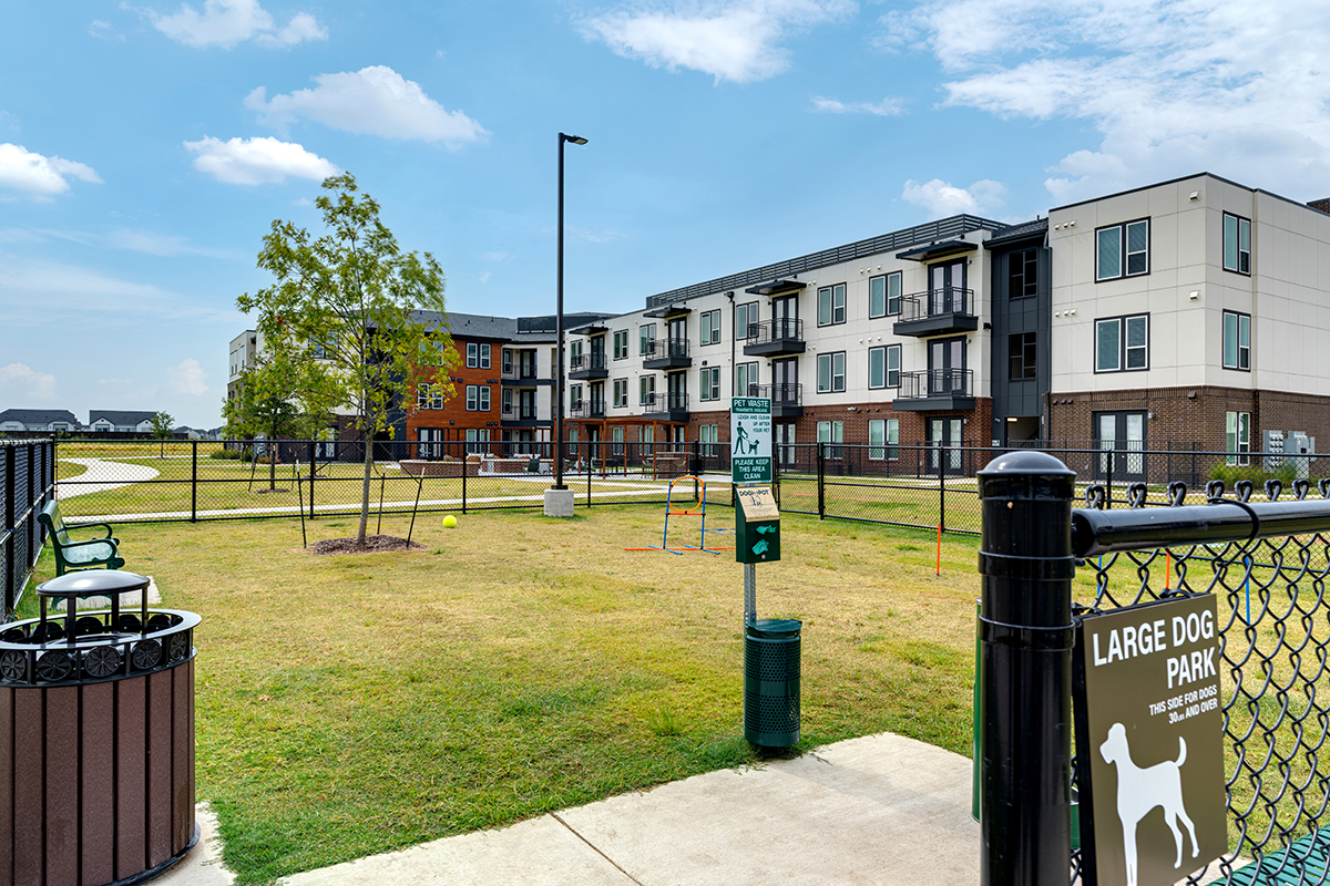 A fenced large dog park with grass, a bench, waste station, and dog park sign in front of a modern, three-story apartment complex under a blue sky with clouds.