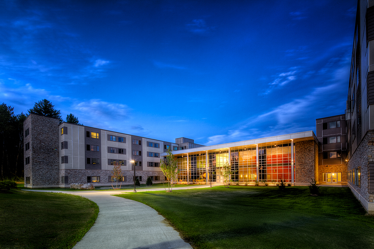 A modern building with large windows and multiple floors is lit up at dusk, surrounded by grass and trees, with a curved sidewalk leading to the entrance under a blue sky.