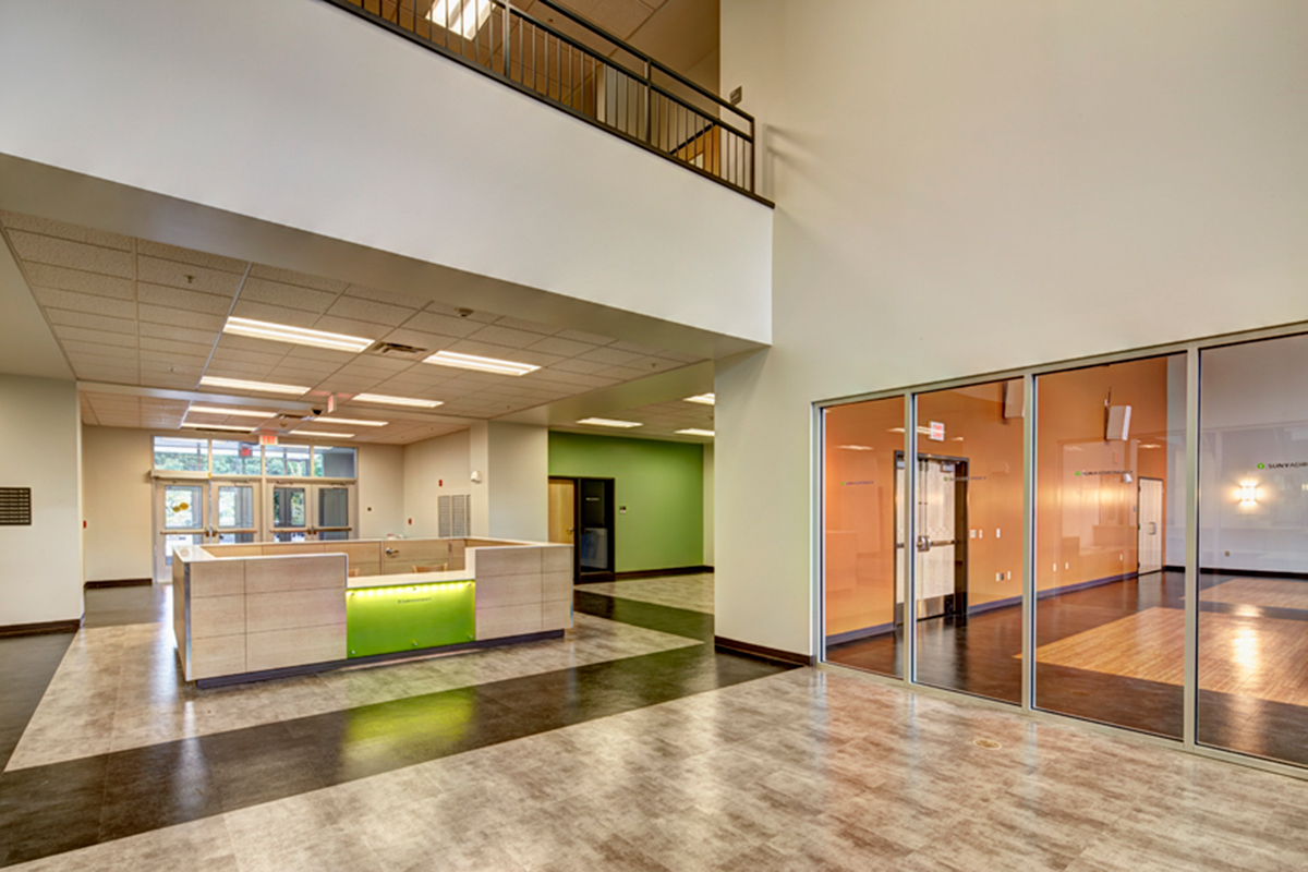 Modern building lobby with a reception desk, large windows, orange accent wall with glass doors, polished floors, and an open second-floor balcony area above.
