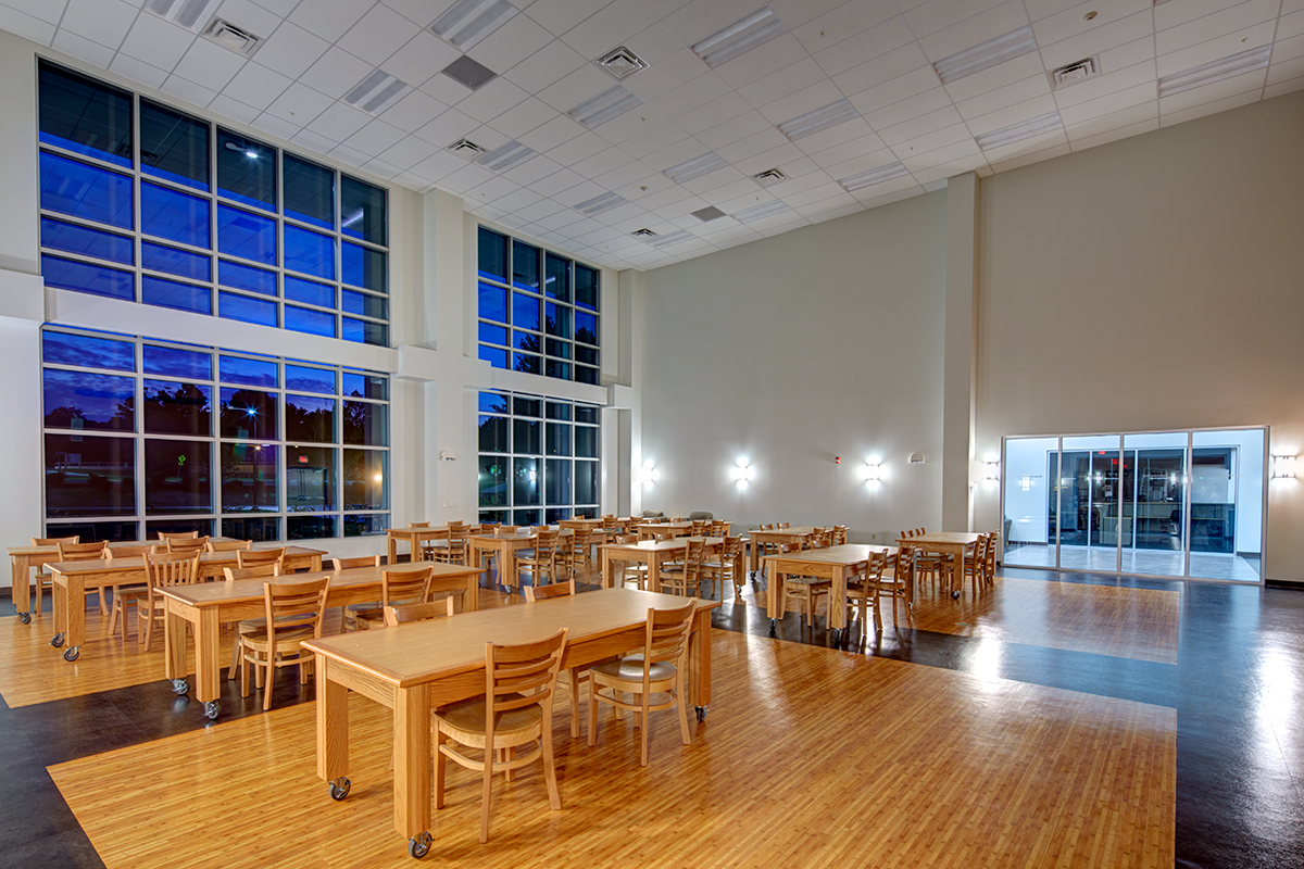 A spacious, modern dining area with wooden tables and chairs, large floor-to-ceiling windows, and a high ceiling. The room is brightly lit with a blue evening sky visible outside.