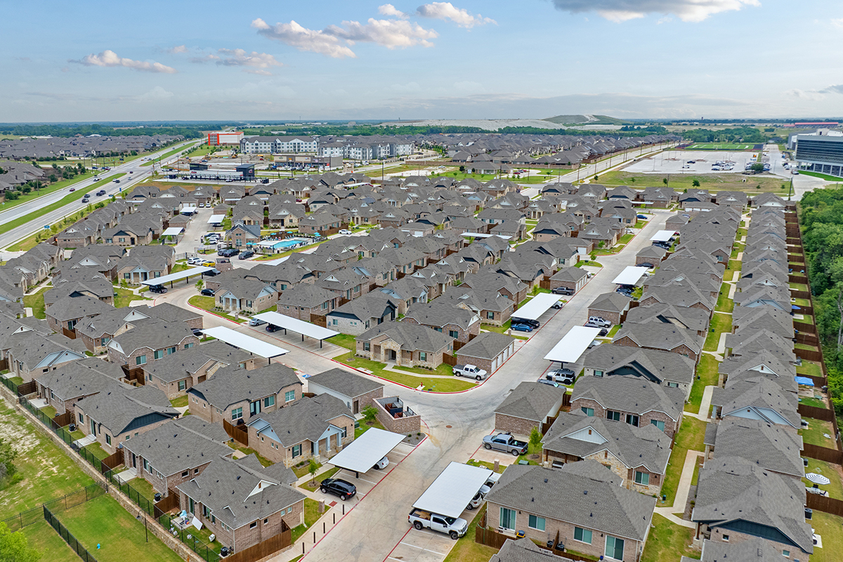 Aerial view of a large suburban neighborhood with rows of similar houses, driveways, and cars, surrounded by roads, parking lots, and green areas under a partly cloudy sky.