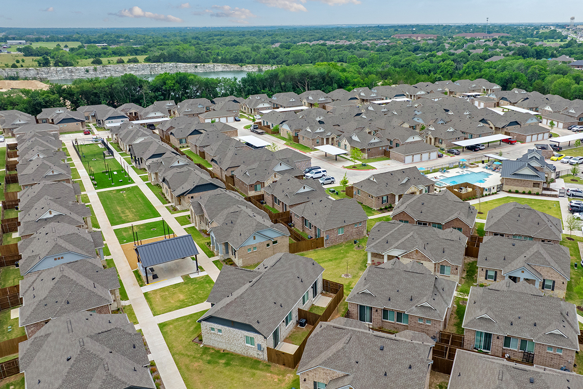 Aerial view of a suburban neighborhood with rows of single-family homes, green lawns, fenced yards, walking paths, and a small park with a covered seating area. Trees and a body of water are visible in the distance.