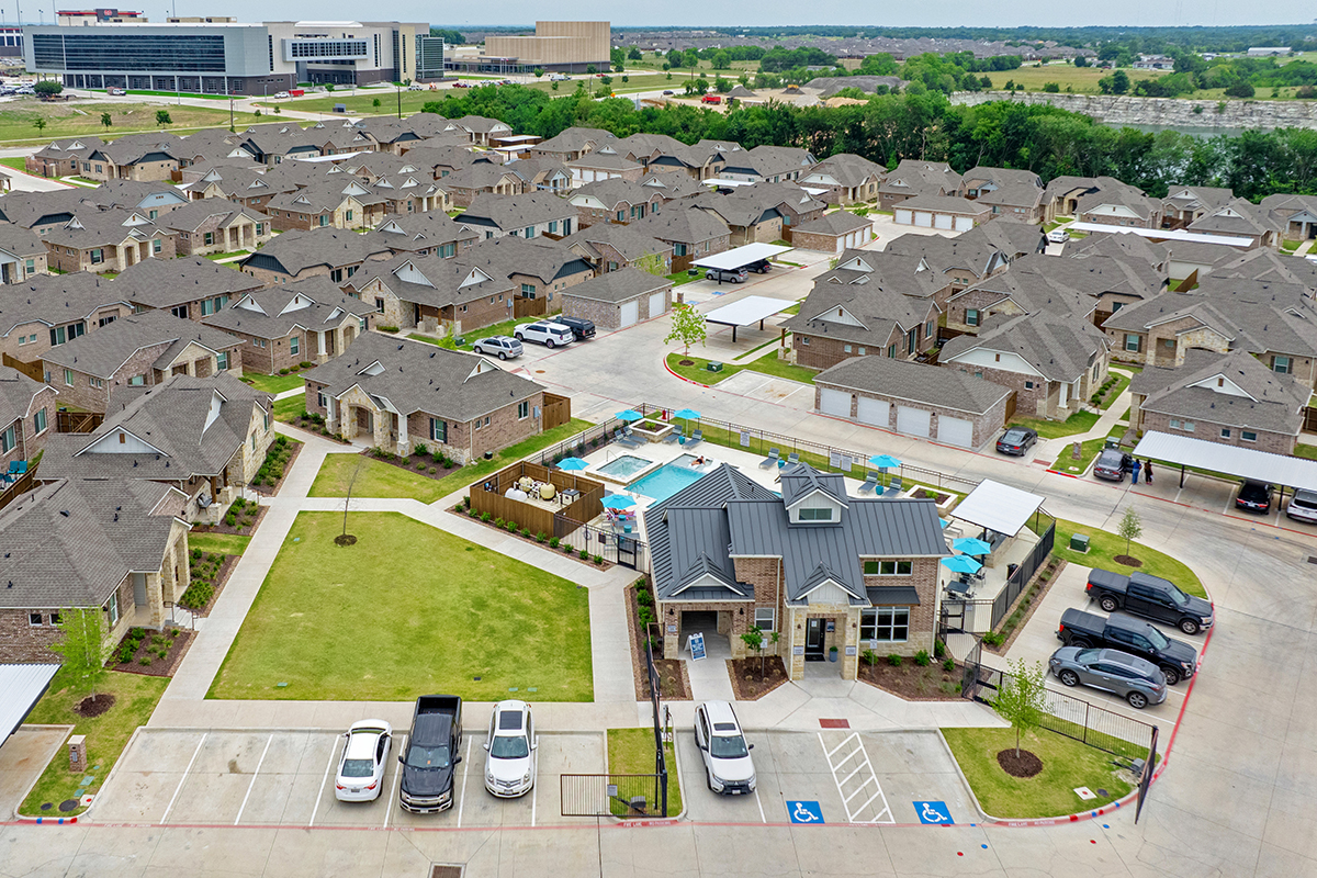 Aerial view of a residential neighborhood with rows of houses, a central community pool, clubhouse, green lawn, parked cars, and nearby streets, set against a backdrop of trees and distant buildings.