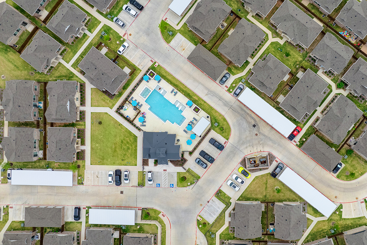 Aerial view of a suburban neighborhood with tan houses, cars parked in driveways, and a central clubhouse area featuring a blue swimming pool with lounge chairs and umbrellas.