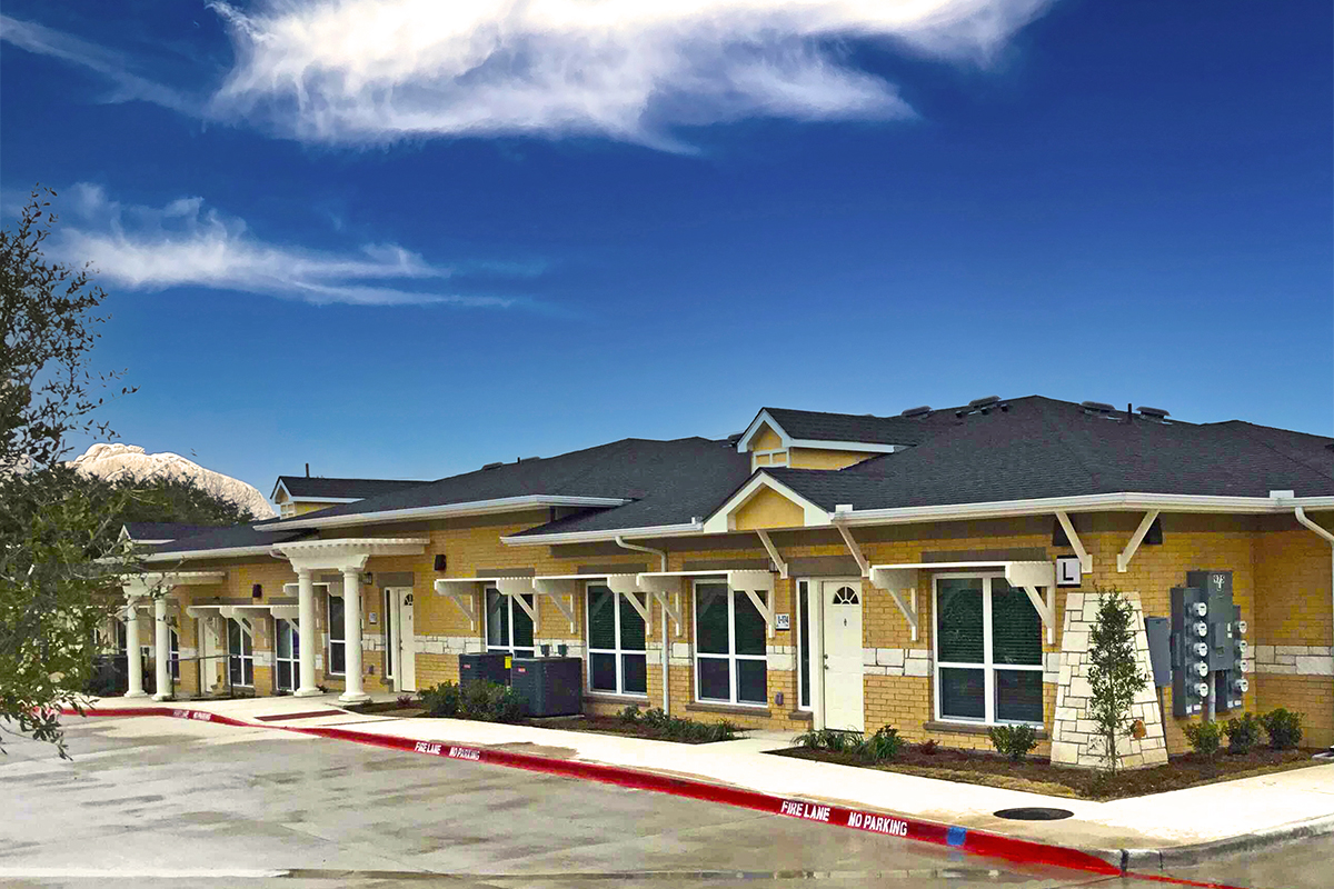Single-story brick building with white trim under a blue sky, featuring several doors and windows. Red curb in front reads FIRE LANE NO PARKING. Small trees and shrubs are planted along the sidewalk.