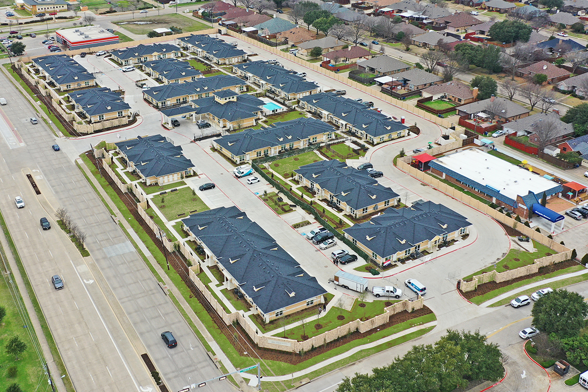 Aerial view of a residential neighborhood with rows of houses, green lawns, parked cars, nearby streets, and some commercial buildings.