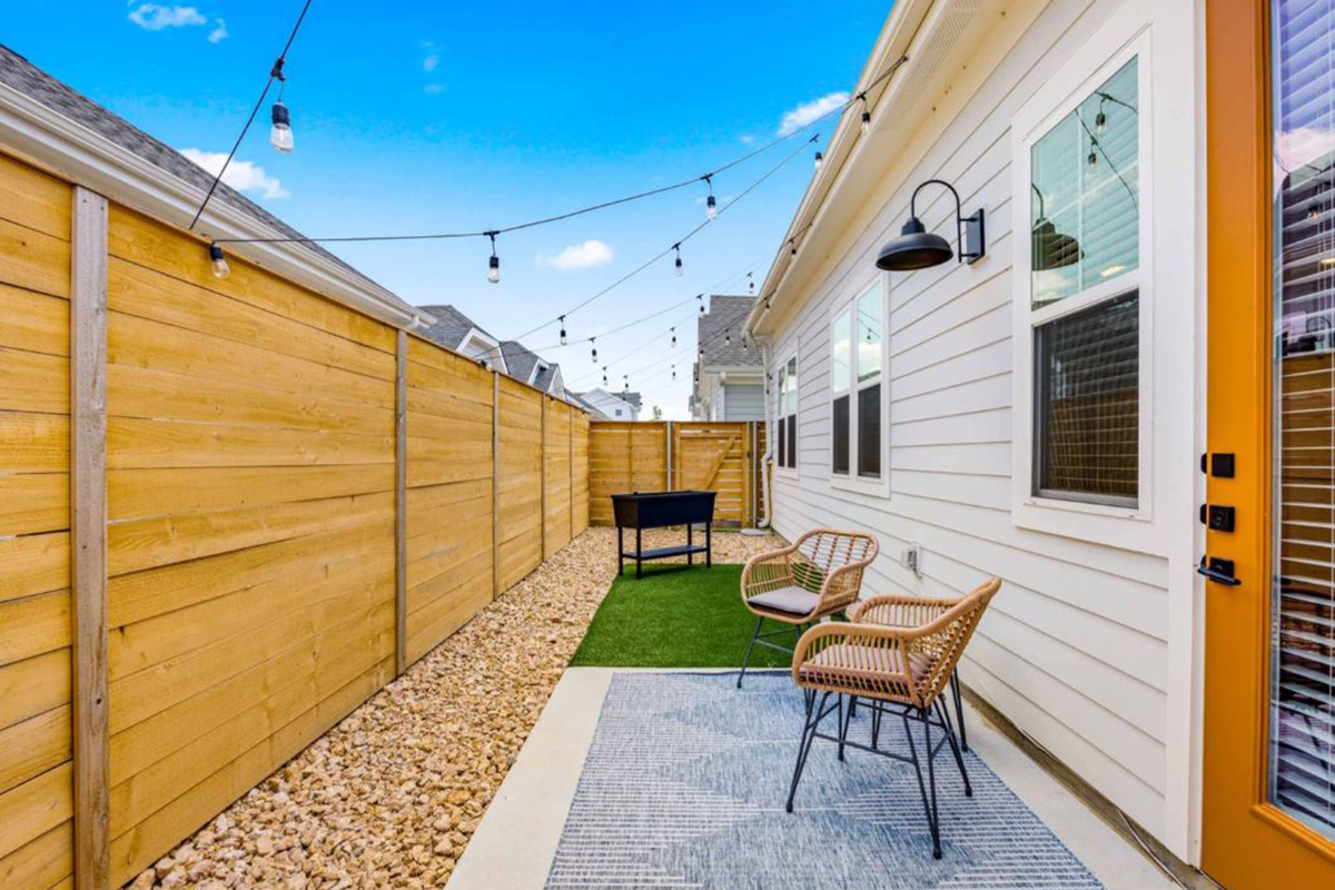 A narrow backyard with string lights, a small grill, two wicker chairs, a rug, and a stone pathway, bordered by a tall wooden fence and a white house with large windows.