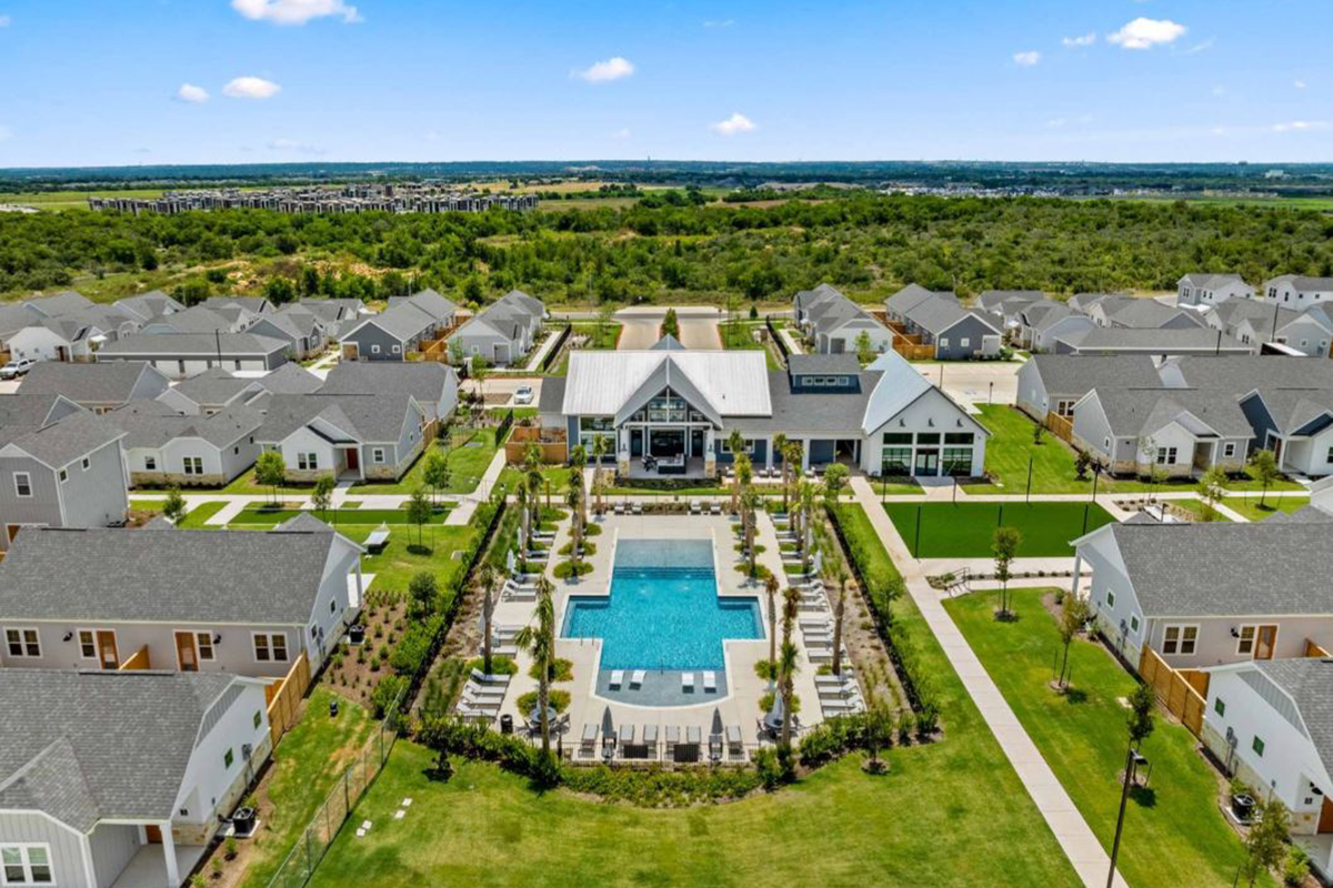 Aerial view of a neighborhood with a central outdoor swimming pool, lounge chairs, landscaped paths, and modern houses surrounding the pool area under a blue sky.