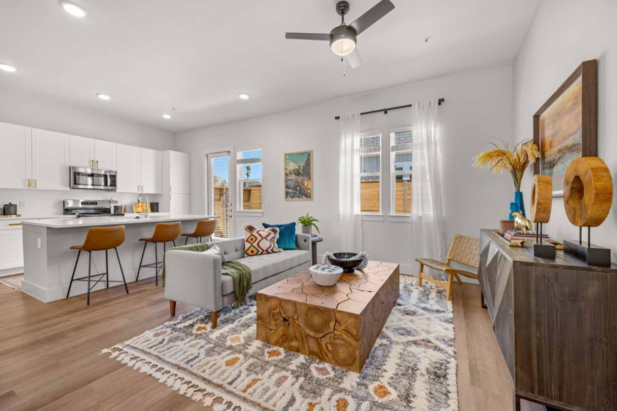 Modern open-concept living room and kitchen with white cabinets, a gray sofa, wooden coffee table, bar stools, rug, artwork, and large windows letting in natural light.