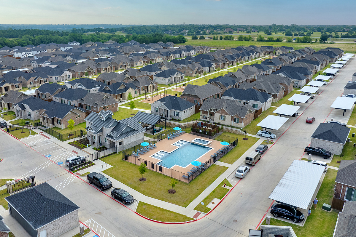 Aerial view of a suburban neighborhood with rows of houses, carports, green lawns, and a central outdoor swimming pool area with lounge chairs and umbrellas.