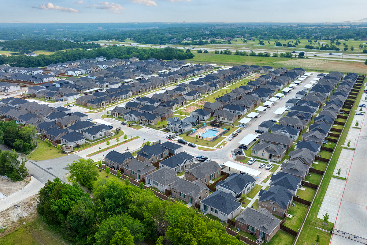 Aerial view of a large suburban neighborhood with rows of similar houses, tree-lined streets, and a central swimming pool, surrounded by open land and a distant highway.