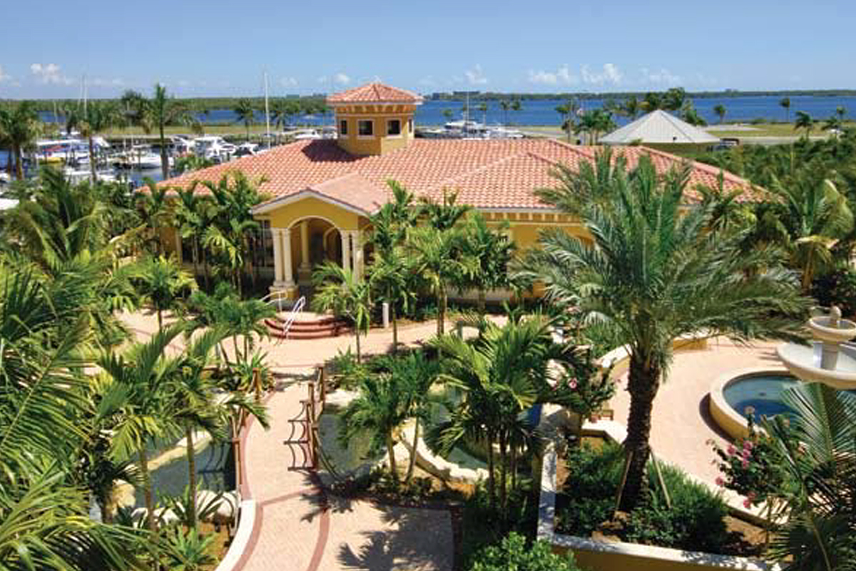 Aerial view of a large Mediterranean-style villa surrounded by palm trees, gardens, and fountains, with a marina and water in the background under a clear blue sky.