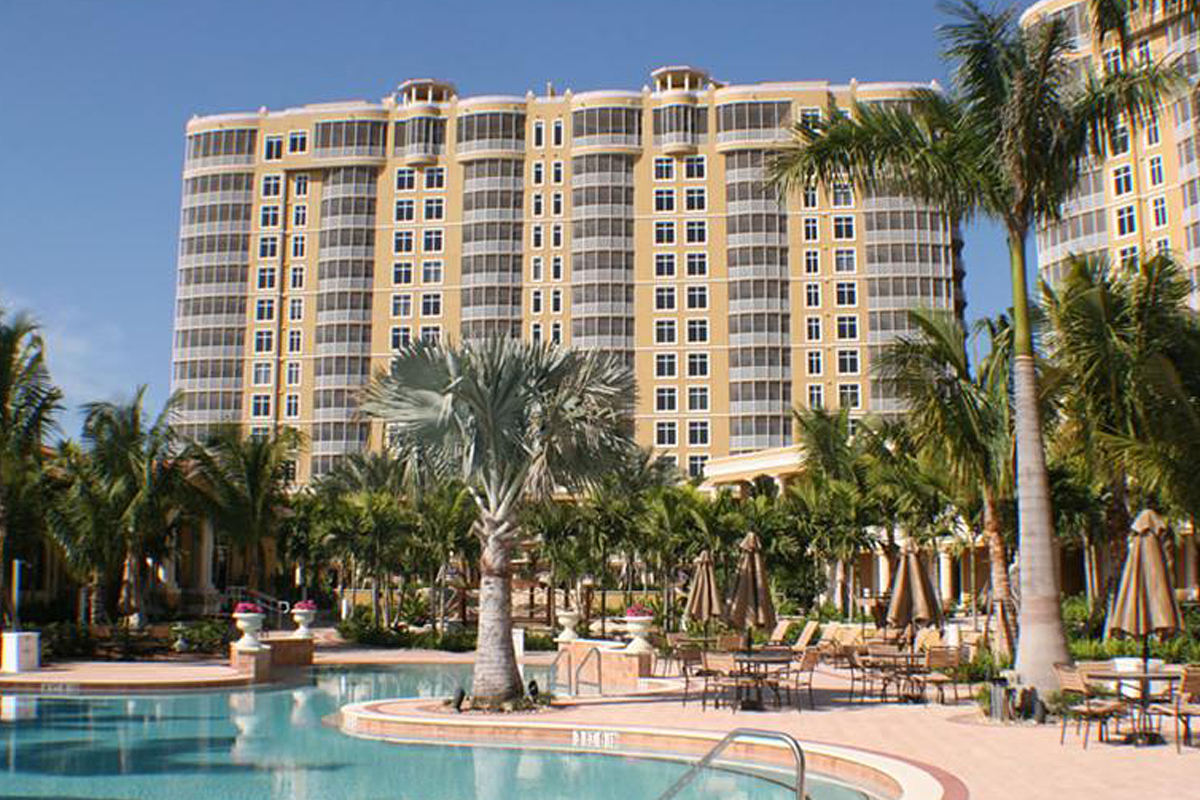 A large beige hotel with many windows stands behind a pool surrounded by palm trees, lounge chairs, and umbrellas under a clear blue sky.