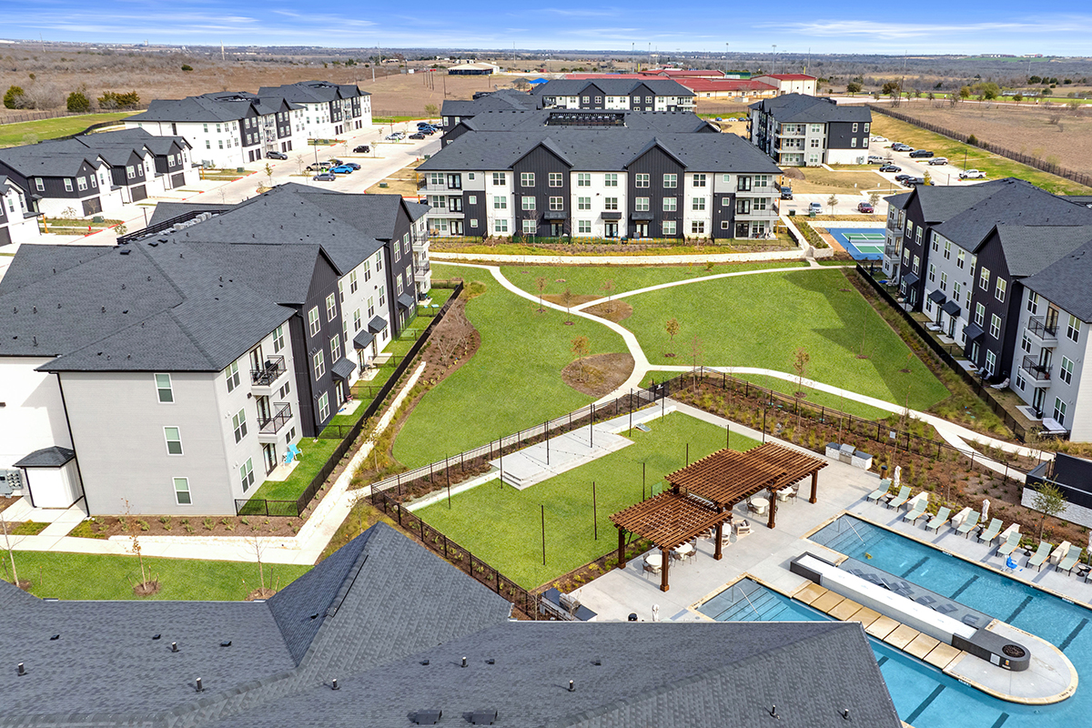 Aerial view of a modern apartment complex featuring several buildings, landscaped green spaces, a pool area with pergolas, and walking paths, surrounded by parking lots and open land.
