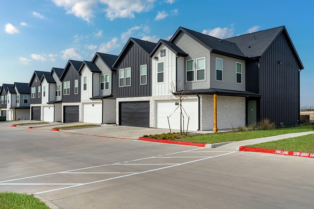 Row of modern two-story townhomes with gray siding, white garages, and small lawns, set along a wide concrete street with clear blue sky above.