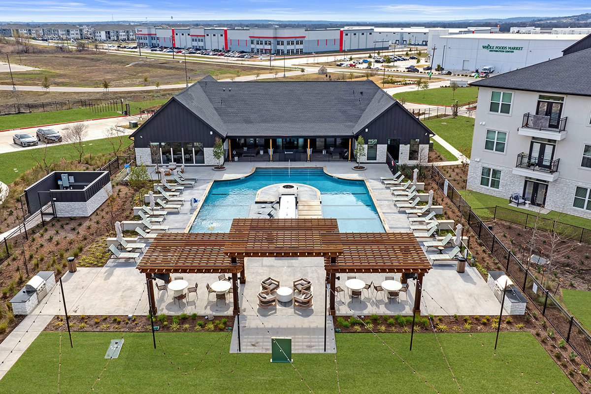 A modern apartment complex pool area with a central pool, lounge chairs, shaded pergola, and seating areas, surrounded by grass, buildings, and a backdrop of industrial warehouses and roads.