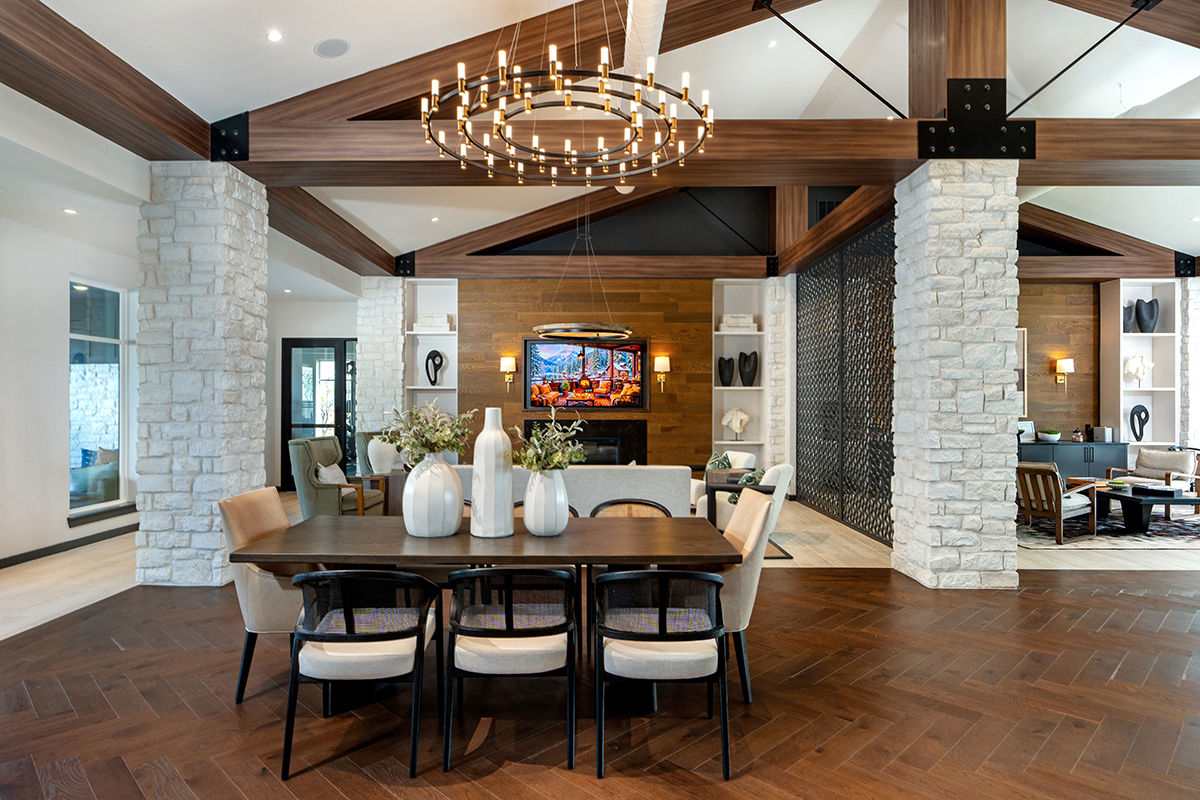 Modern open-concept living space with a dining table, neutral chairs, large chandelier, wood beams, stone columns, hardwood floors, and a TV above a fireplace in the background.