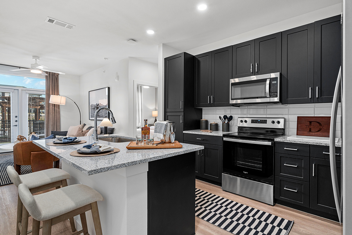 Modern kitchen with black cabinets, stainless steel appliances, a white island with two stools, striped rugs, and a living area with a sofa and large windows in the background.