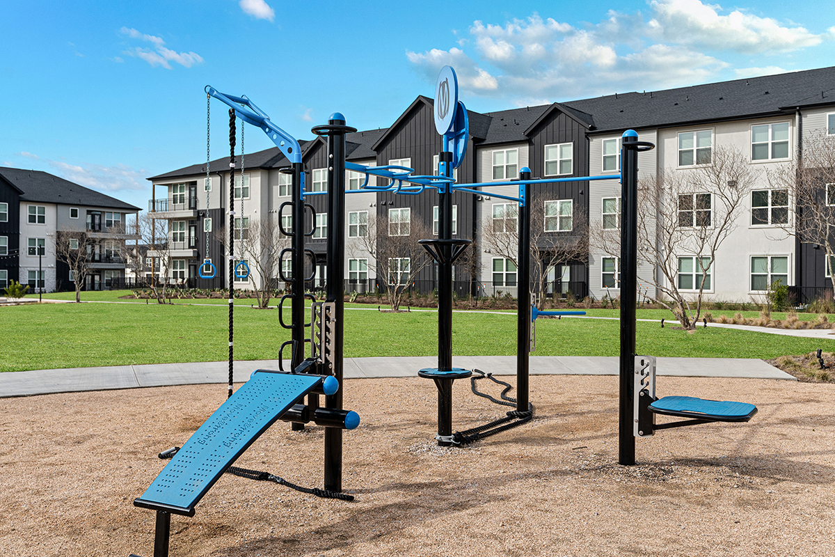 Outdoor fitness equipment set on sand in a grassy apartment courtyard, with modern black and white apartment buildings and blue sky in the background.